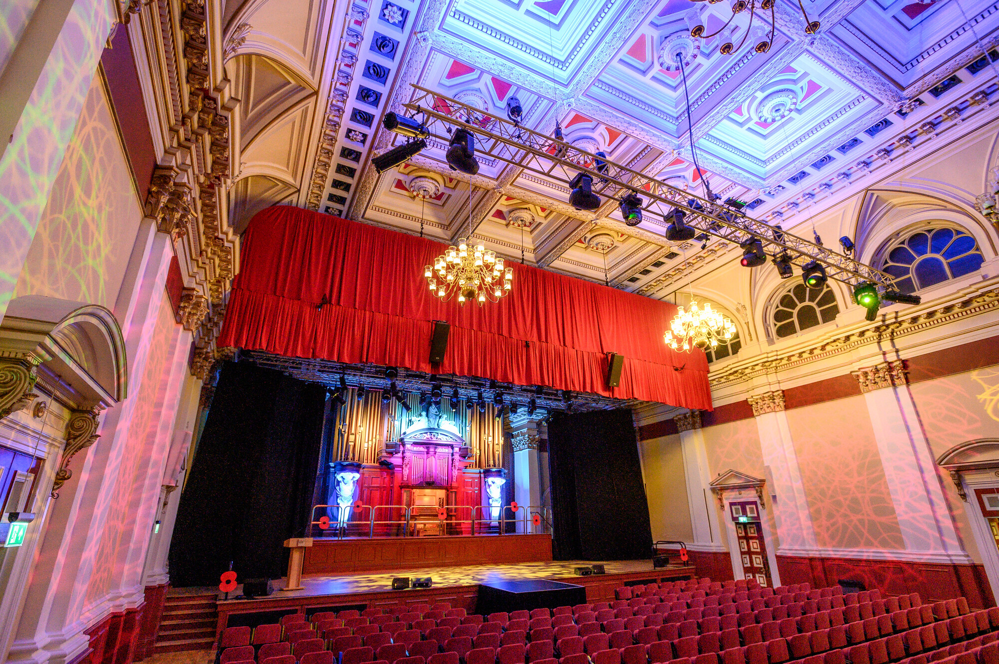 Large ornate theatre hall, highly decorated with large hanging chandeliers. Rows of tiered red seating fills the room facing a stage.