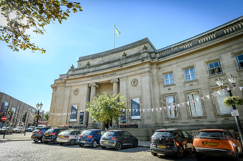 Bolton main library. A stone building with two neo classical pillars. Cars parked out the front. A sunny day with bunting hanging outside.